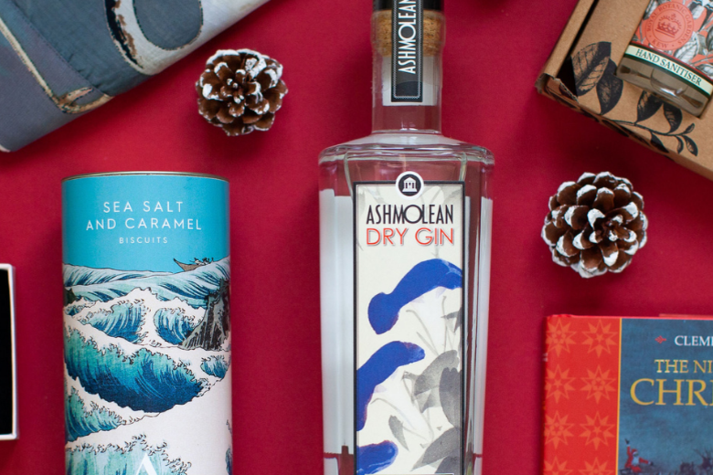 A group of products from the Ashmolean shop against a red background, including a bottle of gin in the centre, biscuits in a blue patterned tin, a book with an orange spine and soaps in orange packaging.