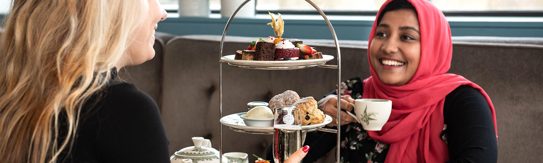 Image of a lady wearing a pink headscarf holding a cup of tea with an afternoon tea stand in front of her