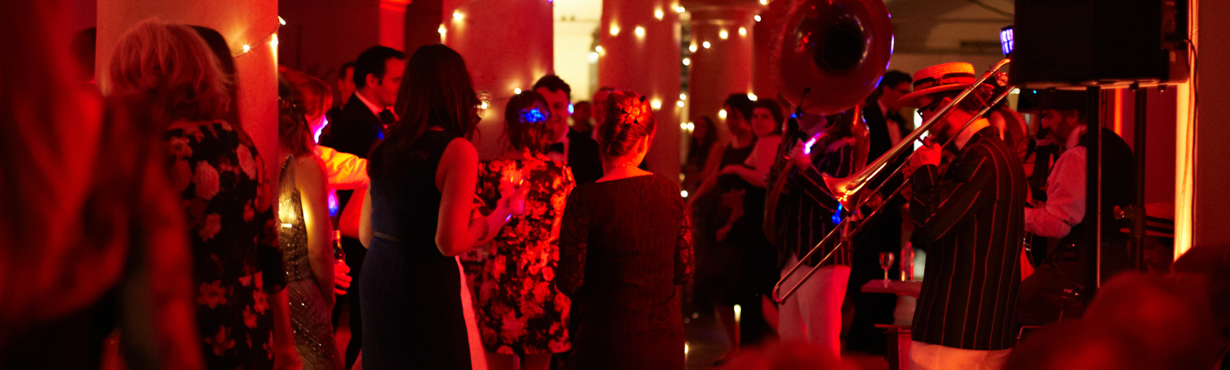 An image of people celebrating Christmas in a vaulted room lit in a red hue with fairy lights adorning pillars and a trombone being played by a musician