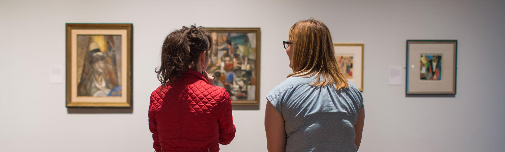 The backs of two women who look at four modern art paintings hung on a neutral wall