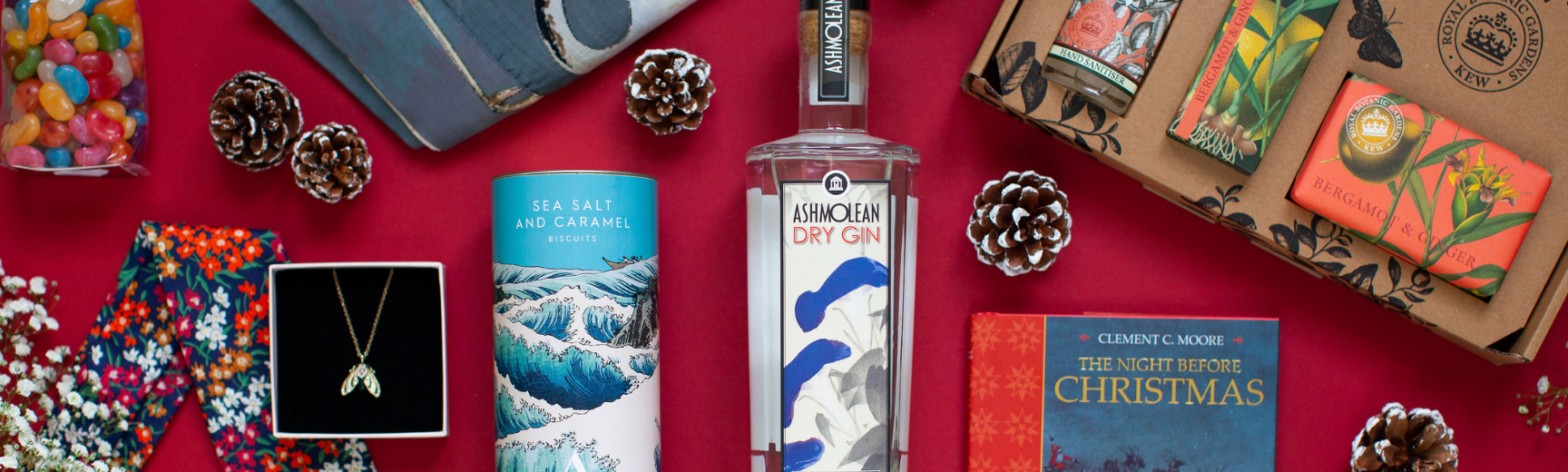 A group of products from the Ashmolean shop against a red background, including a bottle of gin in the centre, biscuits in a blue patterned tin, a book with an orange spine and soaps in orange packaging.