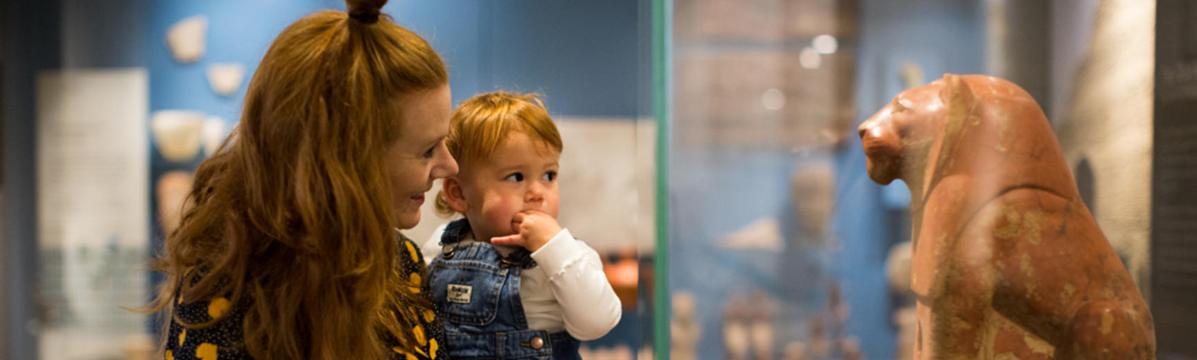 A woman with longer hair holds a small child, and they both look at a red terracotta model of a seated lion inside a glass case