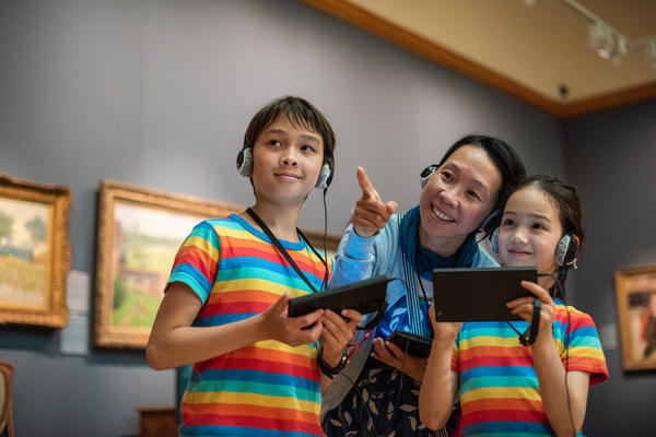 Two children in rainbow t-shirts, and a woman, smile as they use multimedia guides in a museum gallery