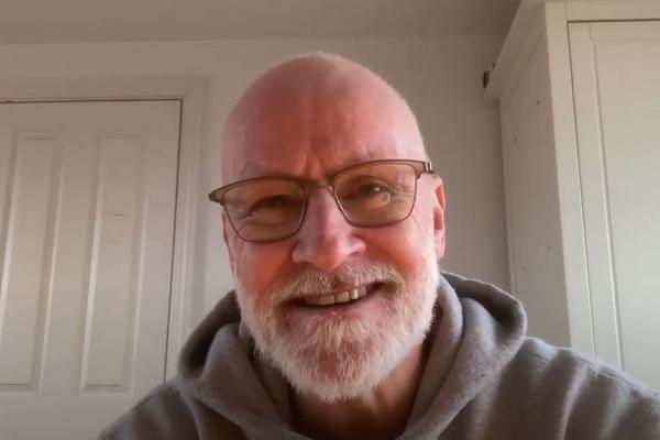 A man with beard and glasses speaks to a webcam, with a white background