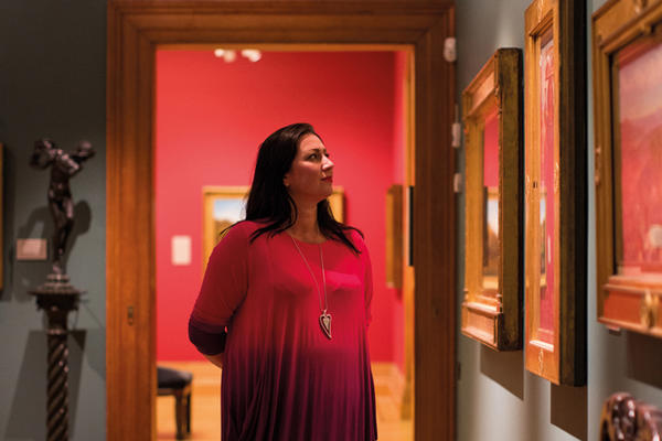 A woman in a red and purple dress stands in a gallery of paintings looking at an artwork