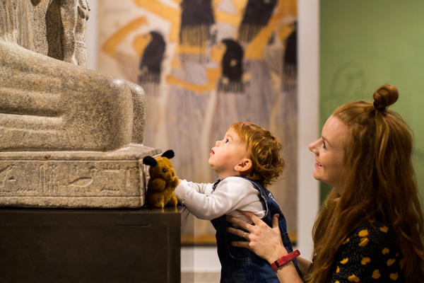 A woman with long hair holds her young child, who wears a white top and denim dress, as he looks up at a stone ancient Egyptian statue