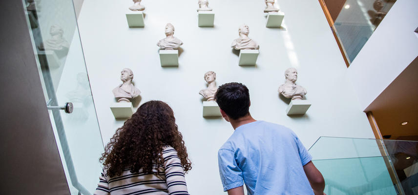 View from behind to museum visitors ascending a staircase, with busts on the wall in front of them