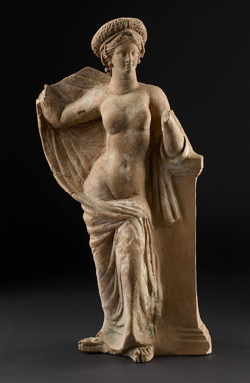 Terracotta figure of Aphrodite posed naked with her hands holding a draped robe around her shoulders and legs
