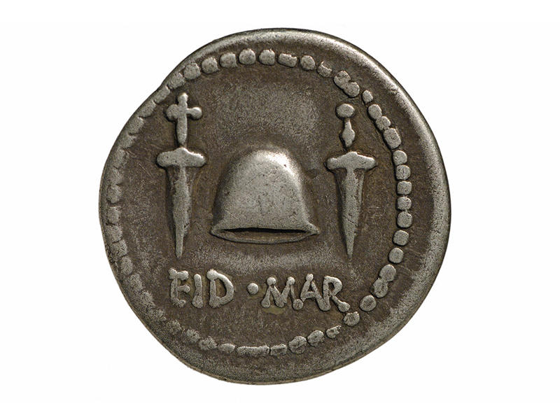 Silver Roman Eid Mar/Ides of March Coin with Daggers and Cap on White Background Silver coin with dotted edge detail, two daggers flanking a centrally position cap, and words EID MAR along bottom edge.